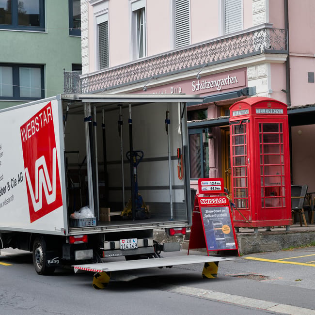A moving van from movingvan.london is parked on a city street in front of a building with pastel-colored facades, one of which has a decorative iron balcony. The van's side door is open, revealing an