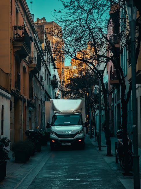 A moving van from movingvan.london is parked on a city street in front of a building with pastel-colored facades, one of which has a decorative iron balcony. The van's side door is open, revealing an