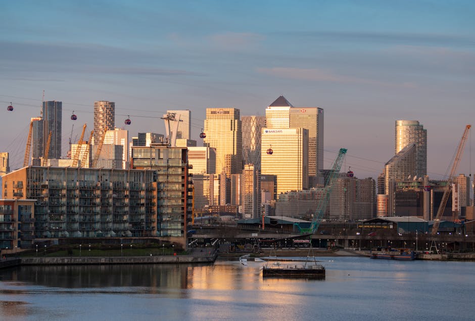 A nighttime cityscape of Canary Wharf in London, viewed across the River Thames. The image shows several illuminated modern skyscrapers with glass facades reflecting city lights, some with red aviatio
