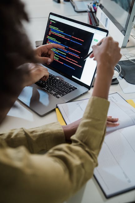 A person seated at a desk inside a property during a house removal process, seen from behind and working on a laptop with a screen displaying colorful lines of code. The desk holds a closed notebook a