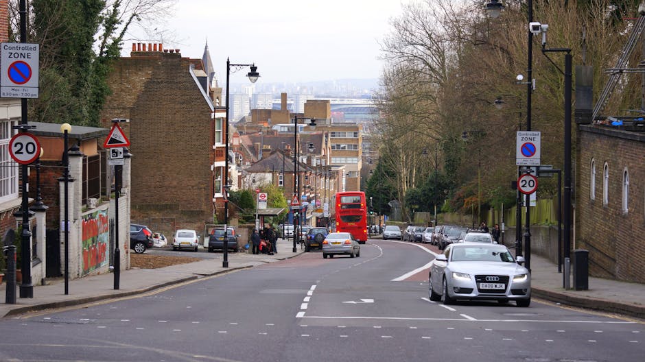 A white and red London Council permit electric shuttle vehicle stationed on a street during daytime, with a man wearing a high-visibility vest visible through the driver's side window. The vehicle bea