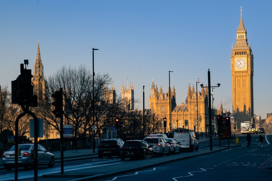A view of Westminster, London, during daylight hours with the prominent Big Ben clock tower and the Houses of Parliament in the background. The foreground features a busy street with multiple cars par