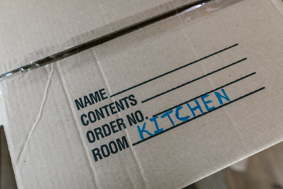 A woman with curly brown hair, wearing a light grey cardigan and white shirt, is seated inside a home, writing on a clipboard with a pen during the packing process for a house relocation. Behind her,